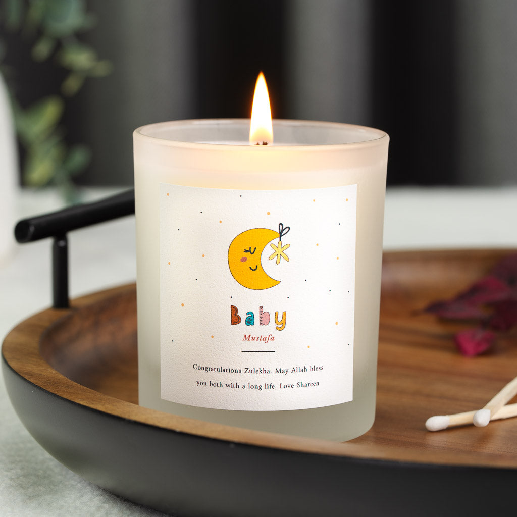 Yellow crescent moon and star above multicoloured type on a personalised new baby candle