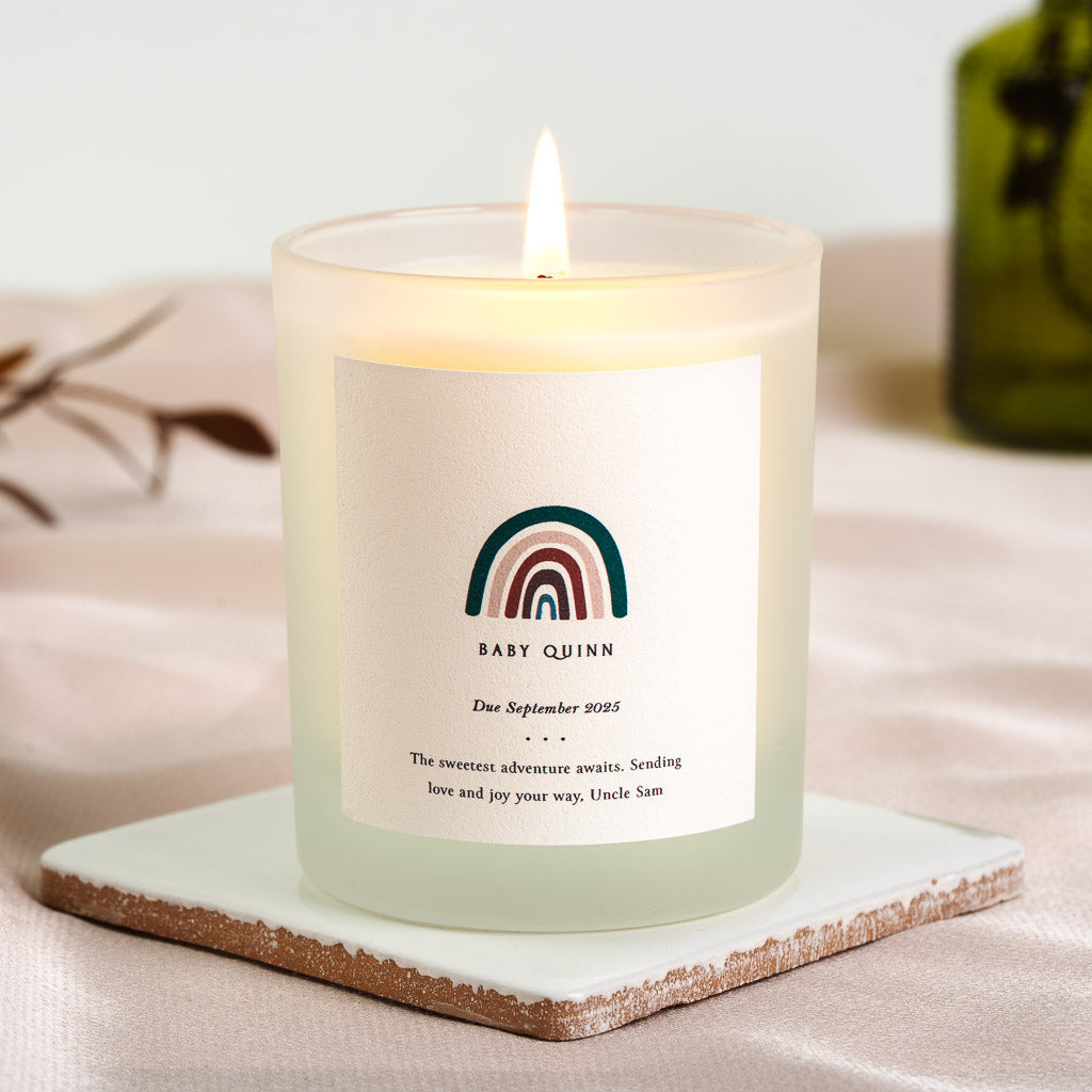 Muted teal, blush and burgundy rainbow above the family name on a personalised baby candle