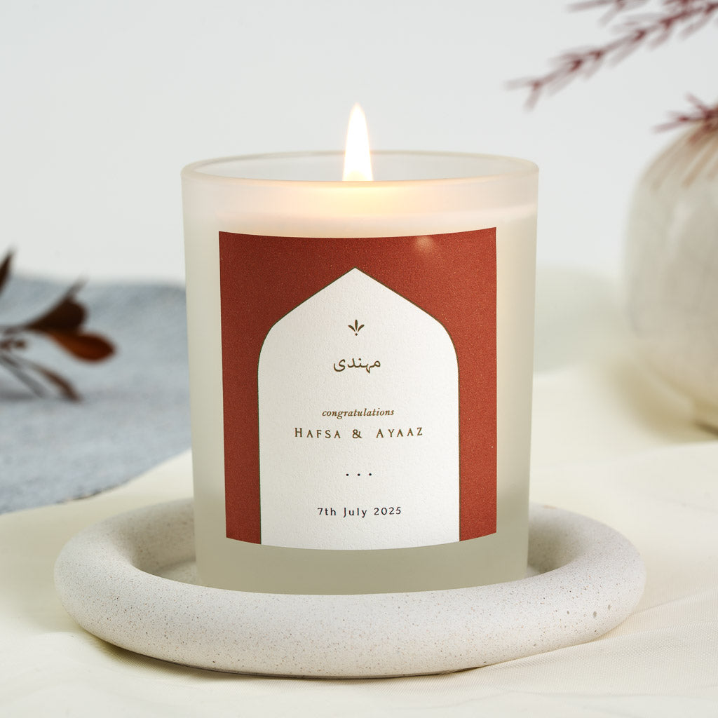 Personalised mehndi candle with gold-toned script inside a white arch on rust red