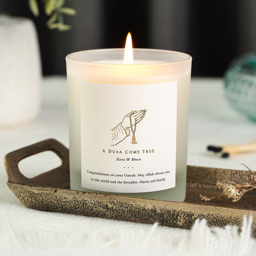 Gold line hand and prayer beads above a duaa line on a personalised Umrah candle label